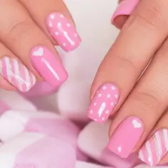 Uroczy manicure hybrydowy na krótkich paznokciach: różowe z groszkami, serduszkami i złotymi paskami, na tle pianek.