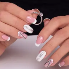 Dłonie z eleganckim manicure, gdzie francuski manicure zdobią kryształki na paznokcie. Delikatny róż i biel tworzą subtelny wzór.