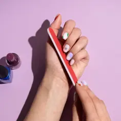Dłoń z kolorowymi paznokciami używa pilniczka. Obok stoją buteleczki z lakierami. Idealne **formy na paznokcie** do domowego manicure.