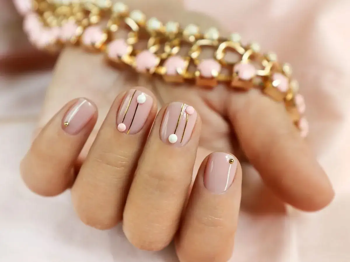 Delikatny manicure hybrydowy na krótkich paznokciach z subtelnymi złotymi zdobieniami i koralikami.