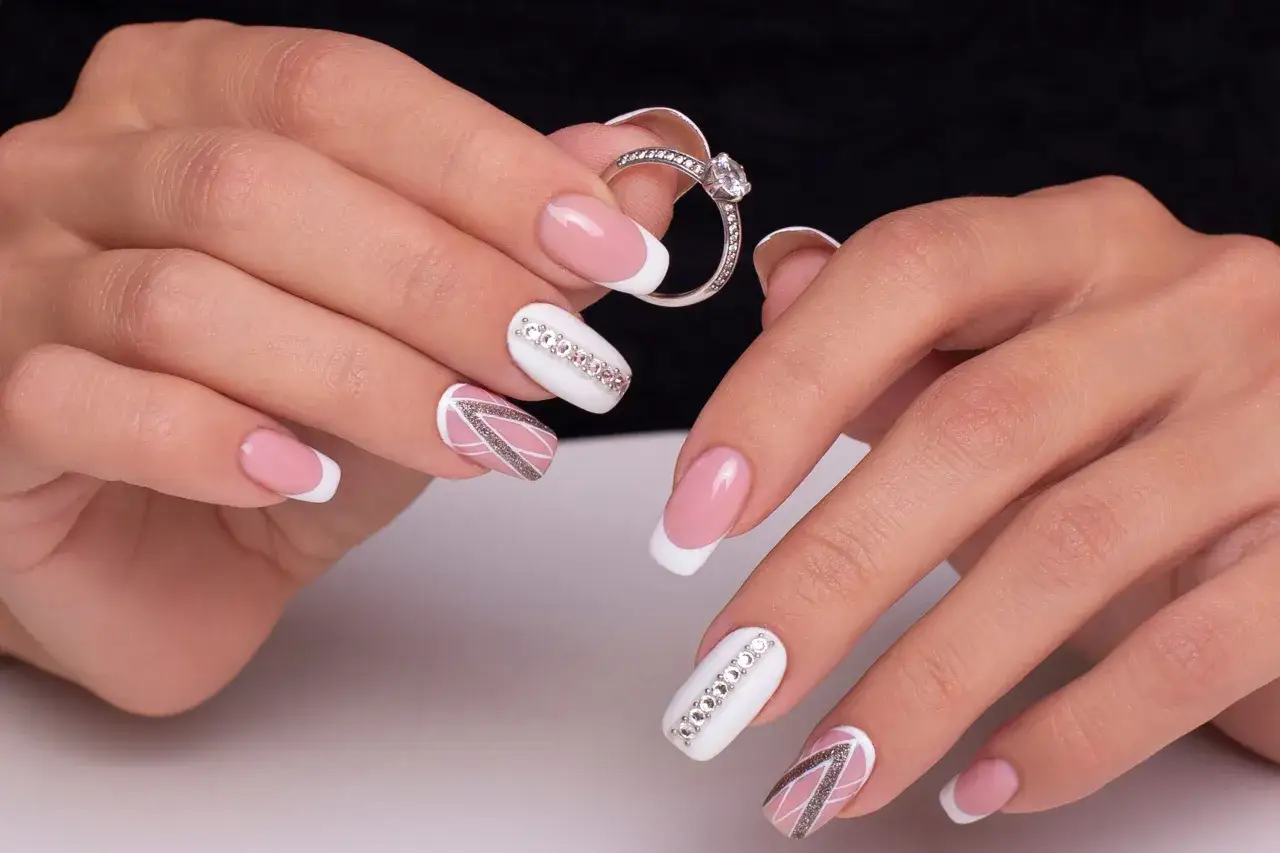 Dłonie z eleganckim manicure, gdzie białe paznokcie zdobią kryształki na paznokcie i subtelne wzory. W tle pierścionek zaręczynowy.